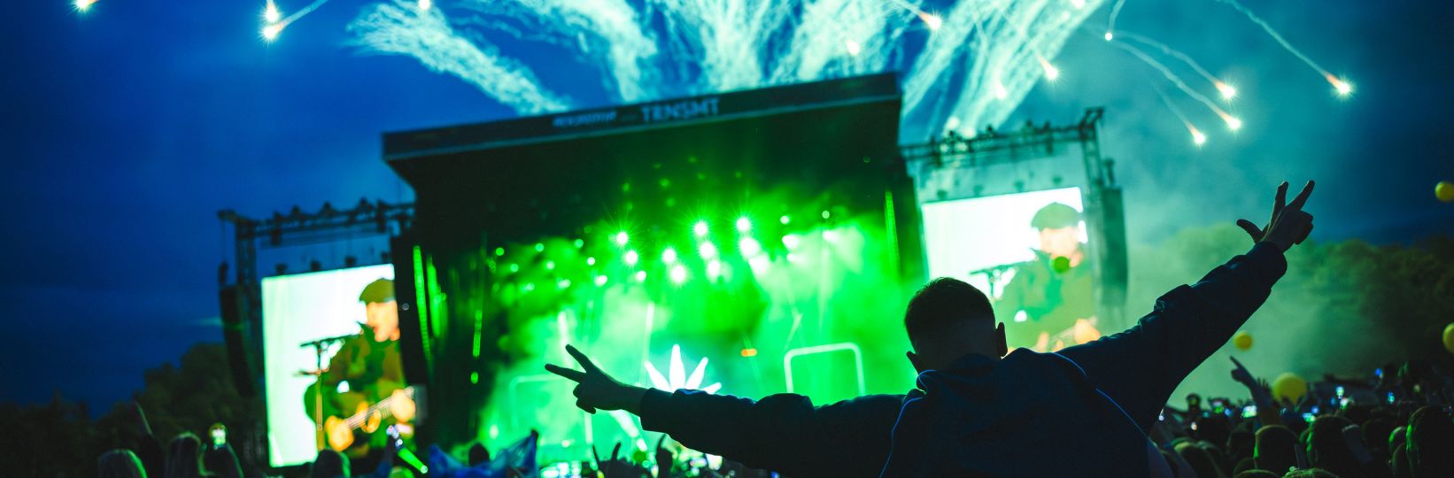 View of Main Stage, green fireworks in the sky and a man on someones shoulders in the audience is in view.