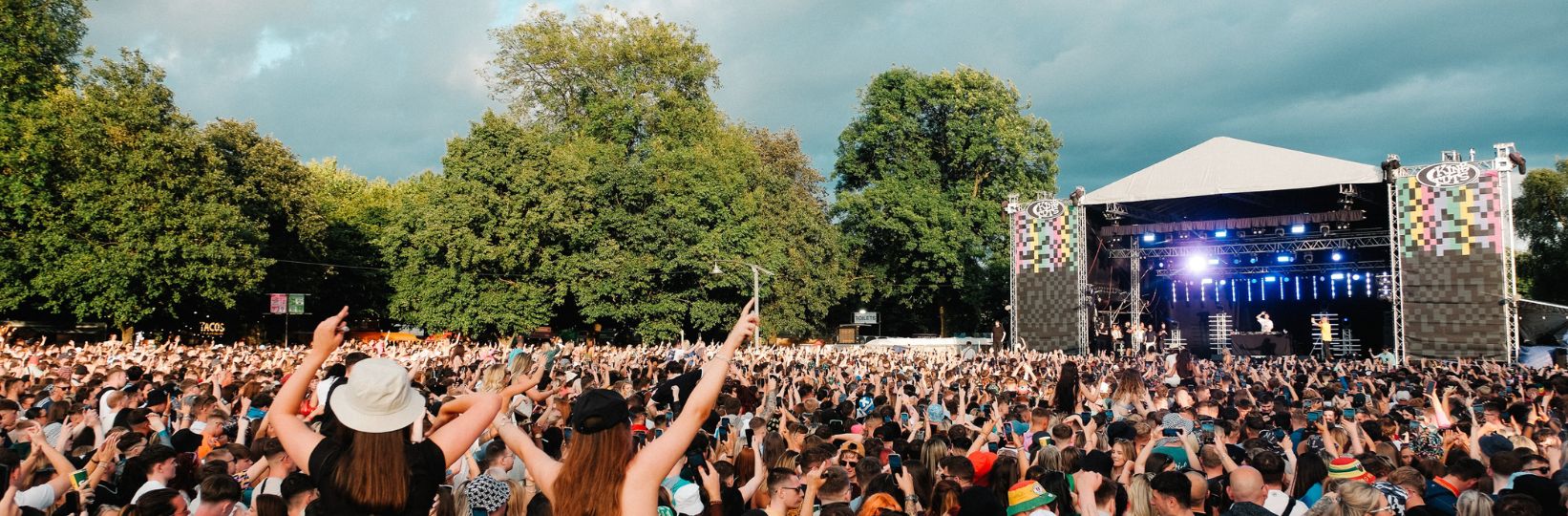 Audience members with their hands in the air, two girls are on shoulders. View of blue sky and trees in background.